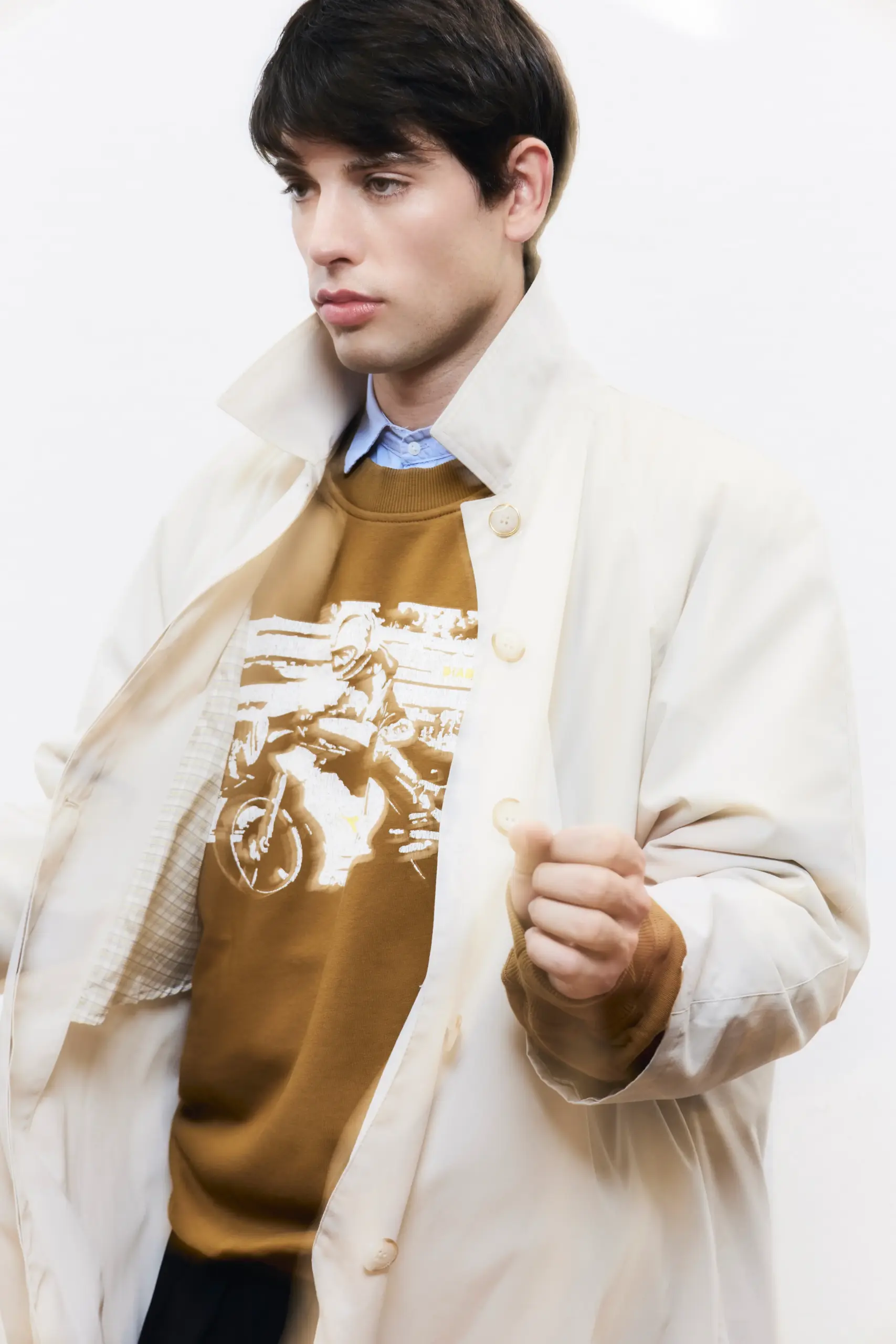 Man in a cream trench coat and mustard motorcycle-print sweatshirt