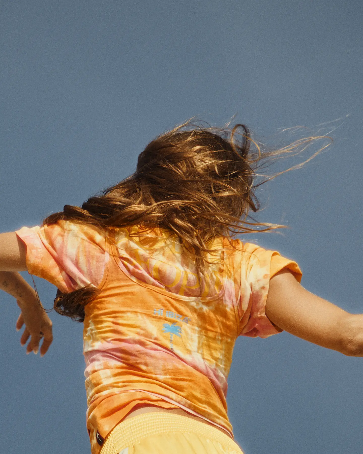 Woman in an orange tie-dye t-shirt, hair blowing, against blue sky