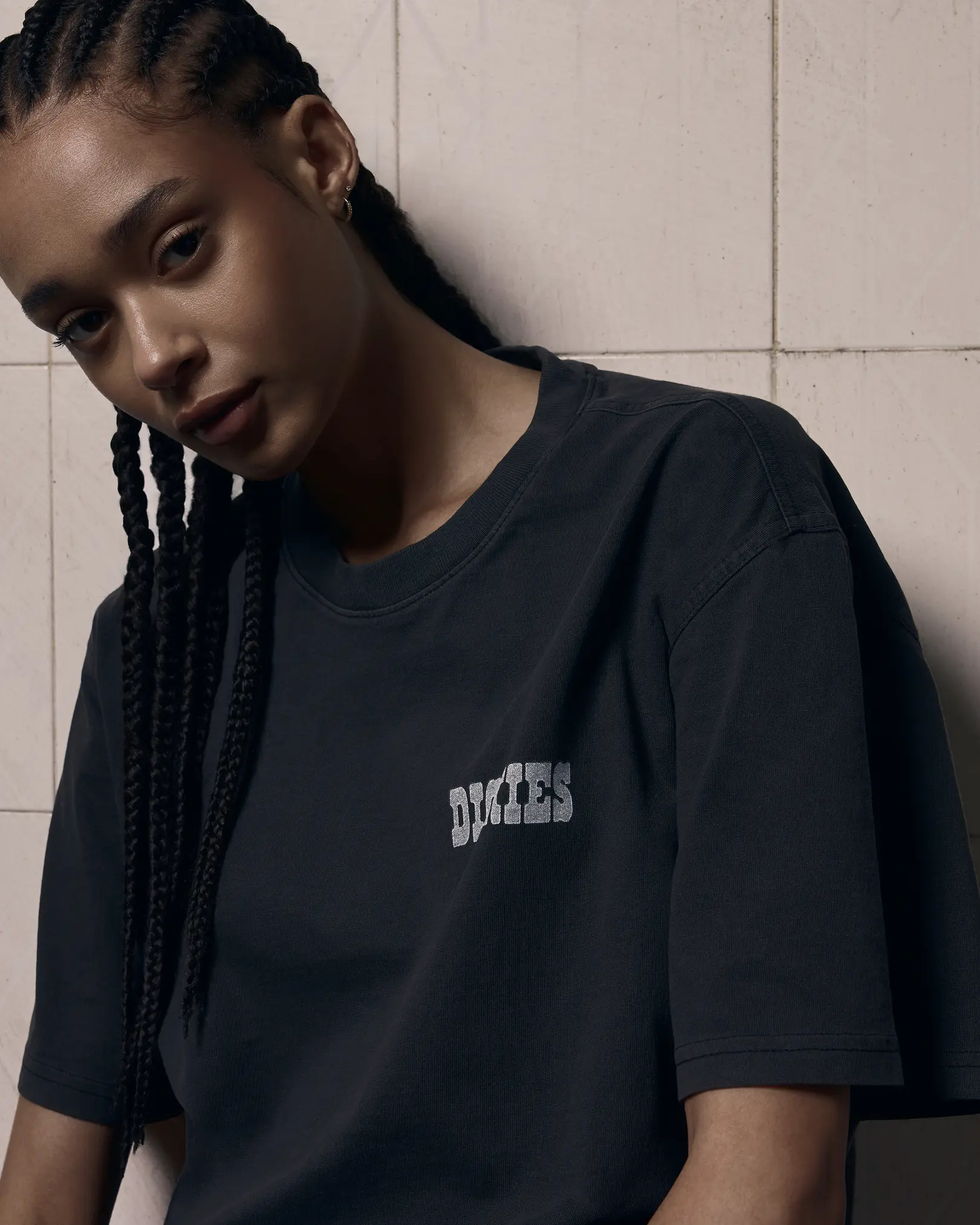 Model with long braids in a black Dickies t-shirt, leaning against a tiled wall