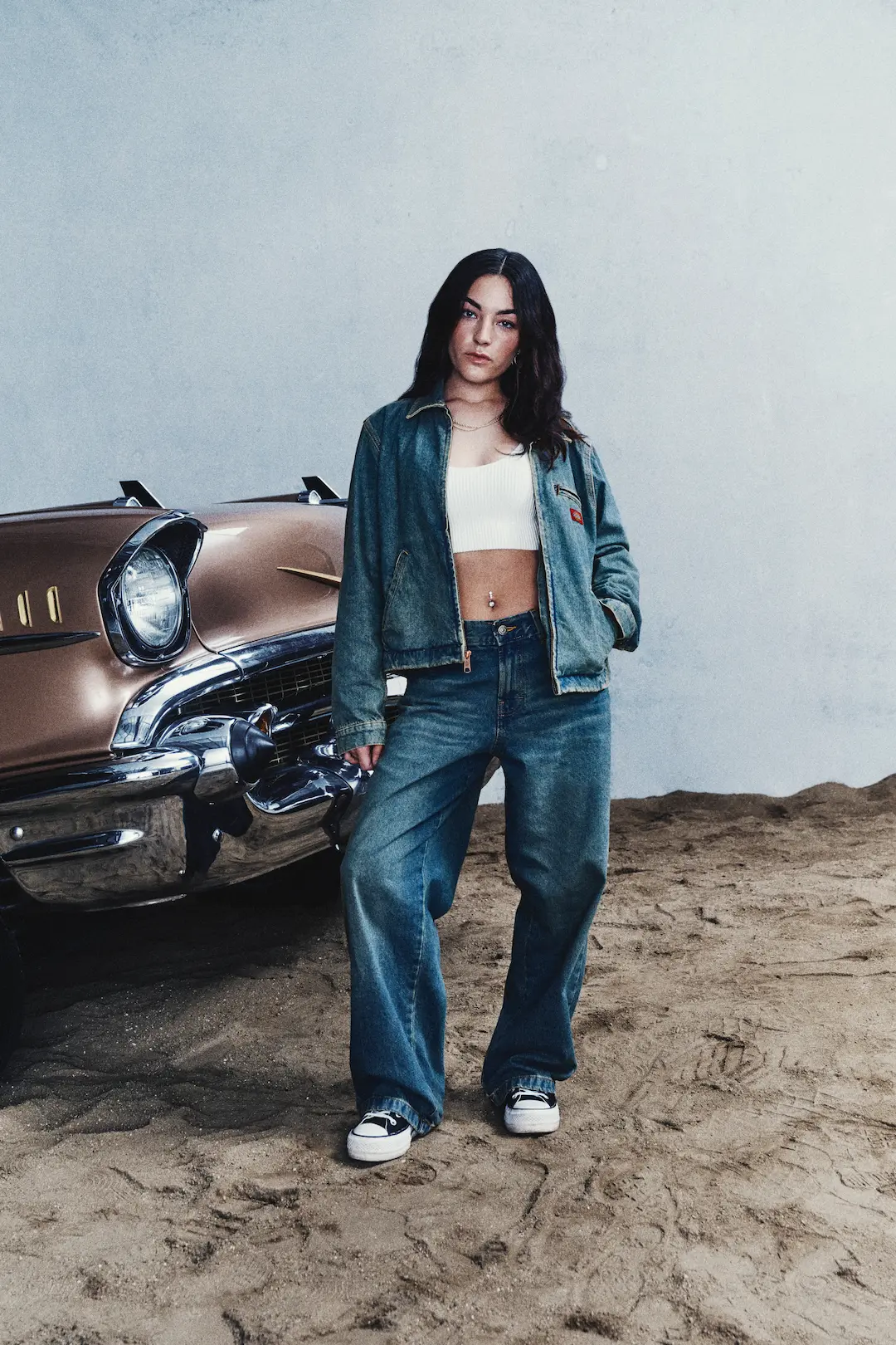 Woman in a denim jacket and baggy jeans beside a vintage car on sand