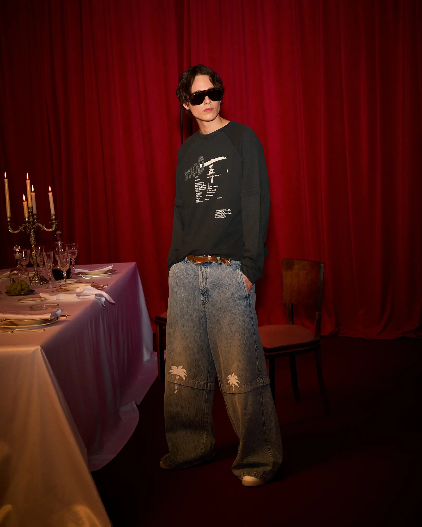 Model in sunglasses and wide-leg Palm Angels jeans before a red curtain