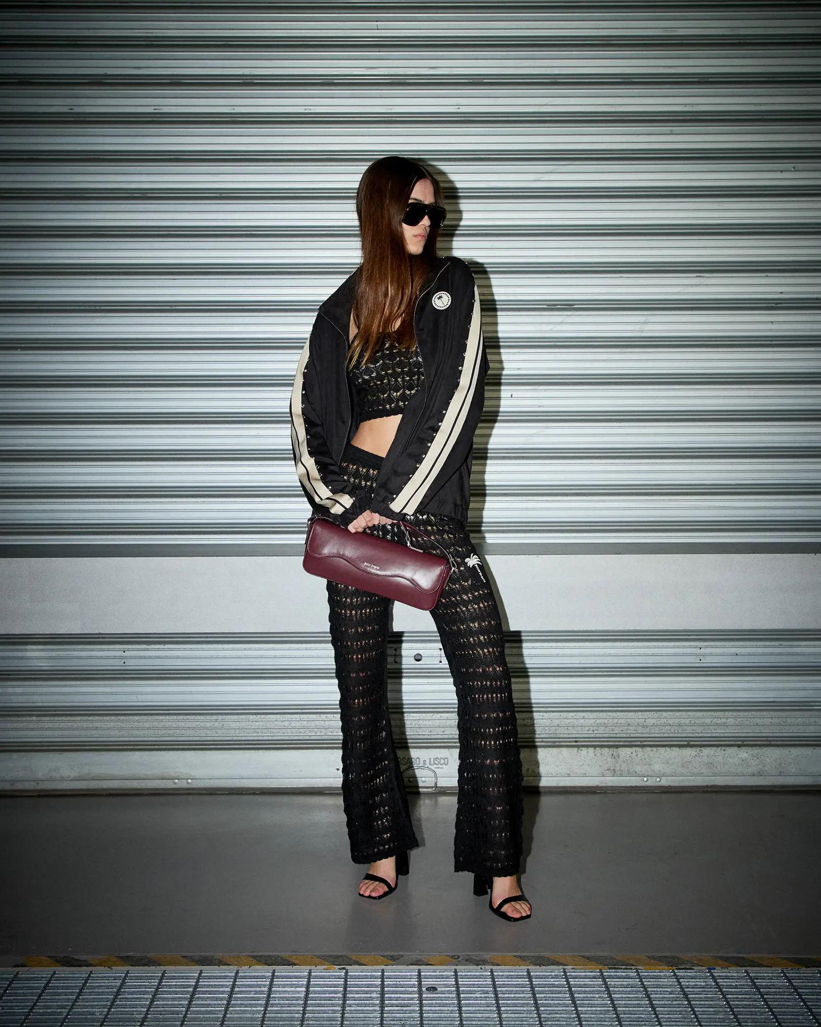 Model in a black track jacket and crochet flares holding a burgundy bag