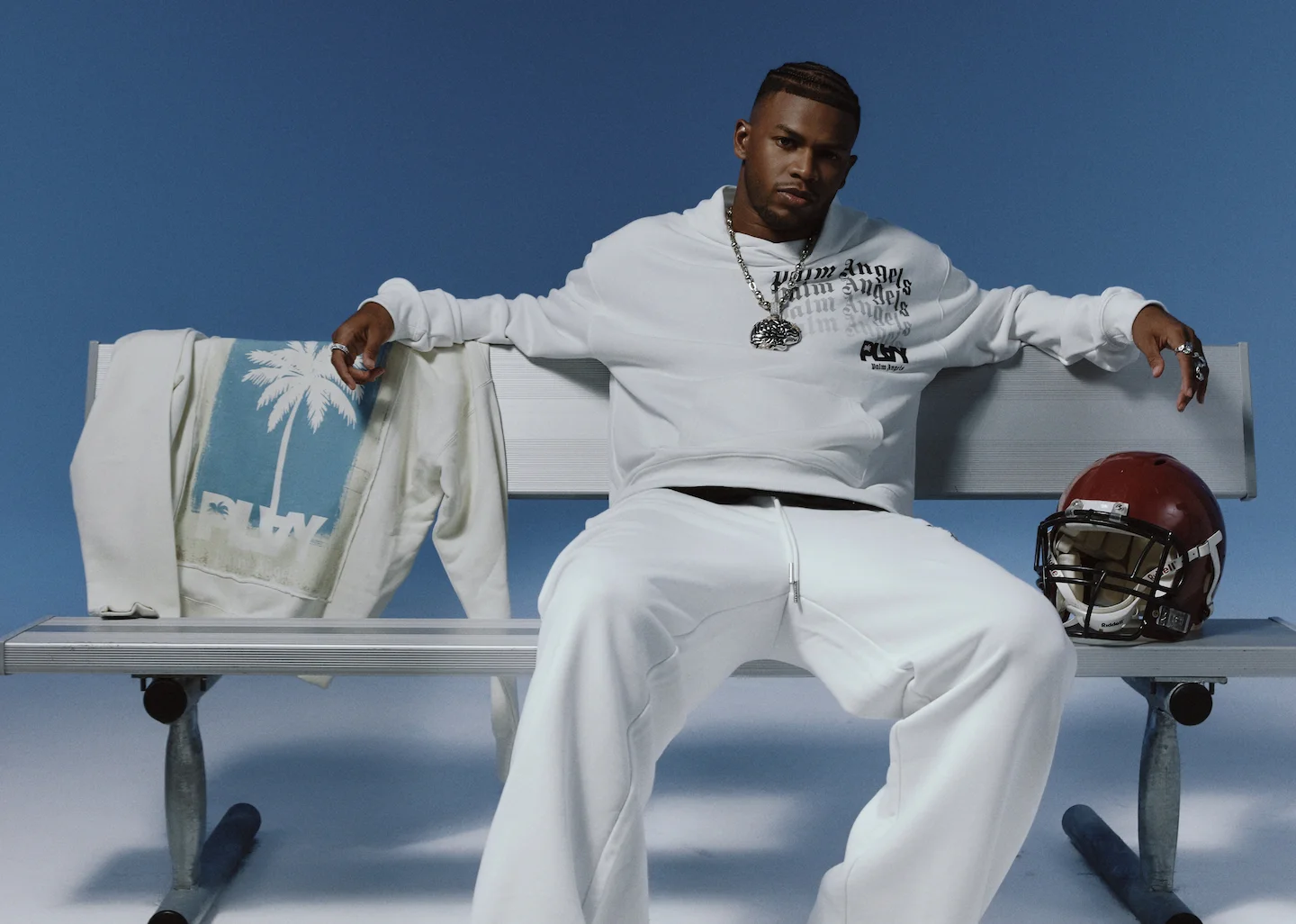 Man in white PLAY Palm Angels loungewear seated on a bench with a helmet