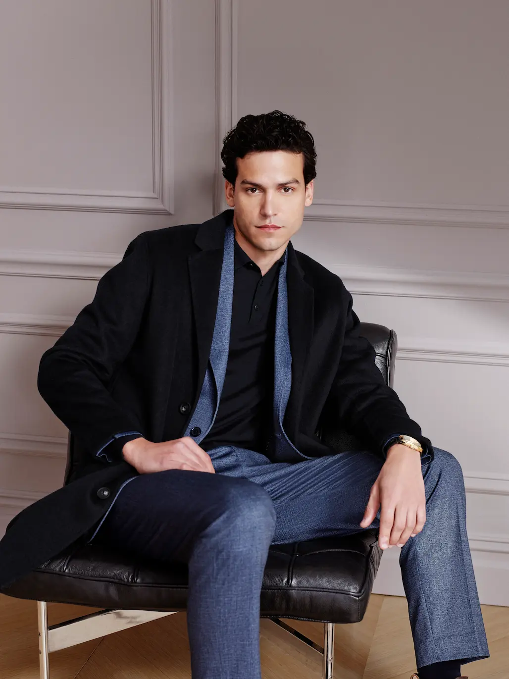 Man in a dark overcoat and black polo seated in a leather chair